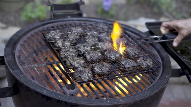 With fire in the grill, a man places a grilling basket full of balls of kofta on the the grill and flames flare up.  Camera moves from left to right.