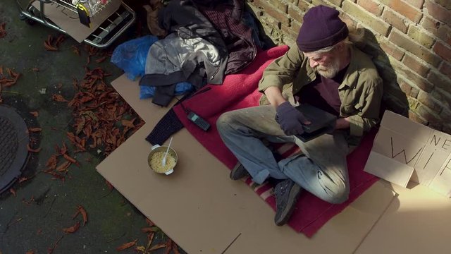 Close up view of homeless old man holding laptop. Tramp sitting on cardboard bu brick wall.