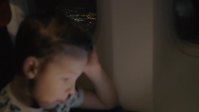Slow motion shot of flying over night city viewed from plane illuminator with following focus on the boy using tablet pc and waiting for landing