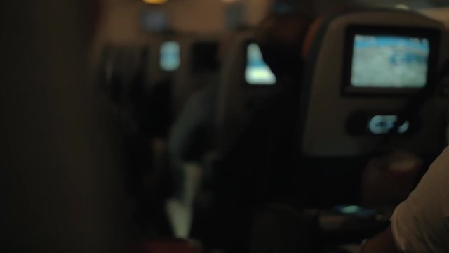 Slow motion shot of passengers following airplane route using the seat monitor map during night flight. View to the cabin with row of seats