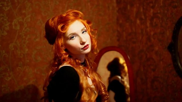Portrait of a woman with long red curly hair in a black and red dress and choker on her neck. Red-haired girl with pale skin, blue eyes, a bright unusual appearance and red lips