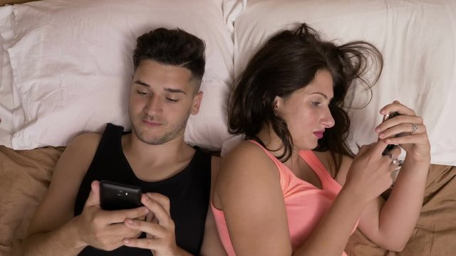 Young couple laying in bed chatting on social media addicted to smartphones ignoring each other
