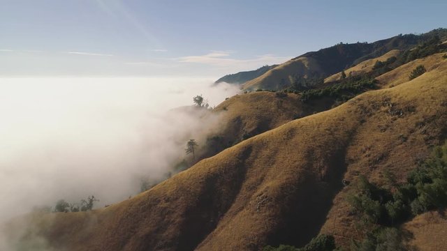 Aerial shot flying forward over fog covered hills. Misty with blue sky above. Fog engulfs the mountains.
