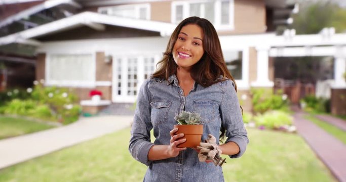 Portrait of attractive Hispanic woman standing on front lawn of home, holding out small plant to the camera. Cheerful Latina female with gardening gloves and equipment smiling at camera. 4k 