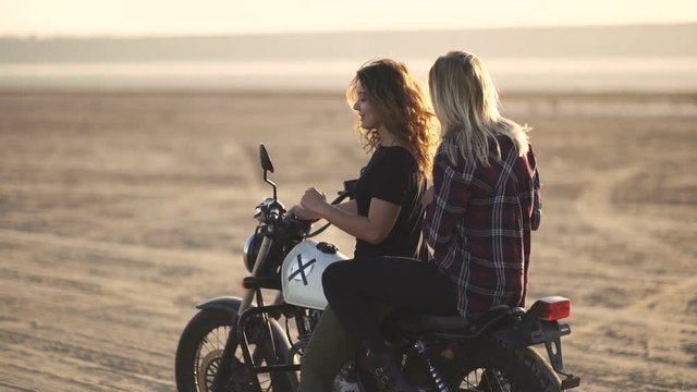 beautiful young woman motorcyclist with his girlfriend riding a motorcycle in a desert on sunset or sunrise. Two woman dancing on a bike