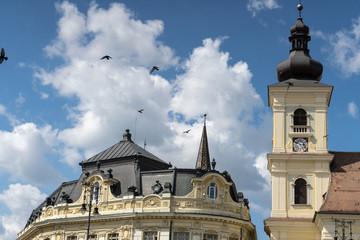 Fototapeta na wymiar Flying pigeons on a cloudy blues sky over the top of buildings in center of Sibiu, Romania