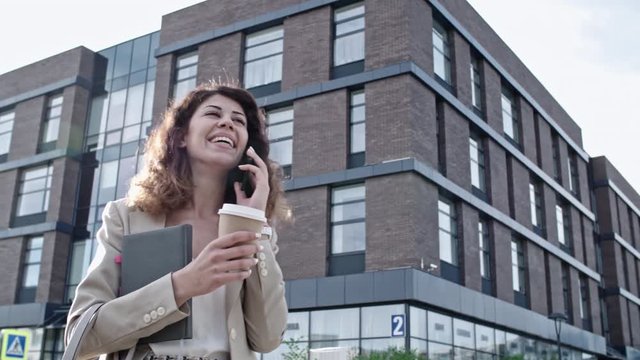 Slow mo of young businesswoman standing with organizer and coffee cup, smiling and chatting on mobile phone at sunny summer day