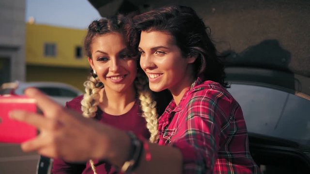 Closeup view of two attractive young women posing while taking selfie on the smartphone sitting inside of the open car trunk. Slowmotion shot