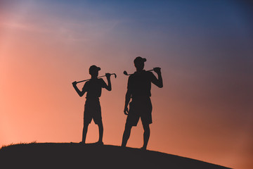 silhouettes of man with his son golfers standing with clubs on golf course at sunset