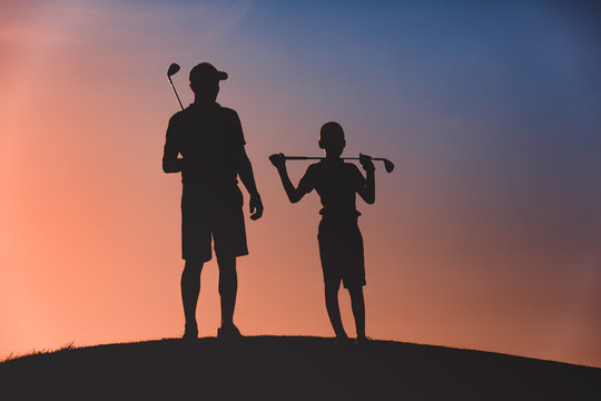 silhouettes of man with his son golfers standing with clubs on golf course at sunset