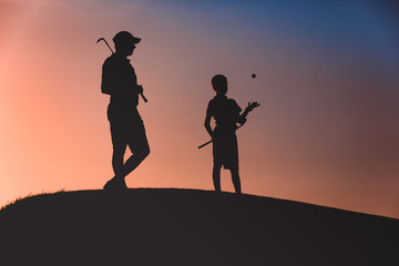 Fototapeta na wymiar silhouettes of man with his son golfers standing with clubs on golf course at sunset