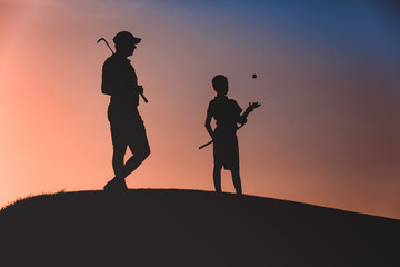 silhouettes of man with his son golfers standing with clubs on golf course at sunset