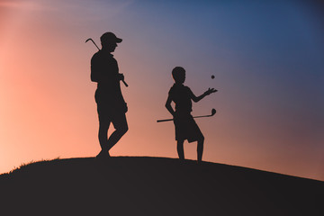 Fototapeta na wymiar silhouettes of man with his son golfers standing with clubs on golf course at sunset