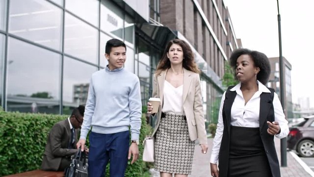 Tilt down of pretty businesswoman holding coffee cup, walking and discussing something with female African colleague and young Asian office worker in downtown
