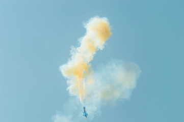Blurry view of an aerobatic plane  leaving a white and yellow smoke trail in the blue sky.