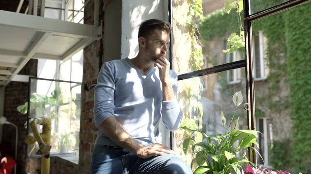 Handsome man sitting on window's sill and looks very worried, steadycam shot
