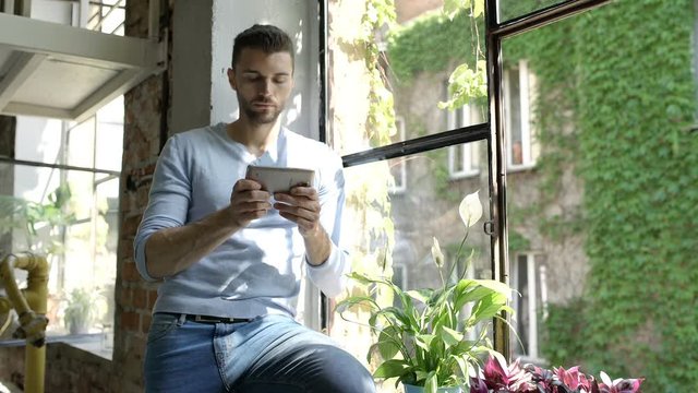 Handsome man in bluew sweater sitting on window's sill and texting messages on smartphone
