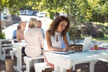 Beautiful woman writing on clipboard while using laptop