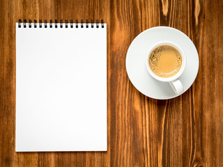 Fototapeta na wymiar coffee in white cup and open notepad with a clean white page on brown wooden table, top view
