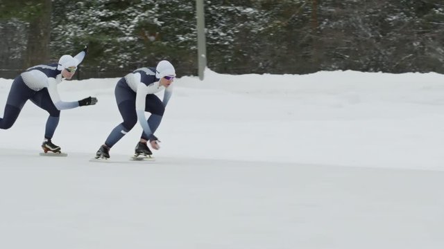 Tracking with slowmo of professional female speed skaters competing against each other in outdoor ice rink in winter; snowed down trees in background