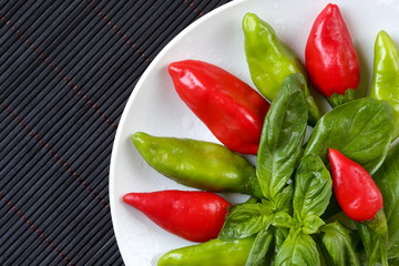 Top view of freshly washed, organic red and green chilli peppers and basil leaves in a white plate on a dark bamboo table mat. Healthy food background with space for text, close up