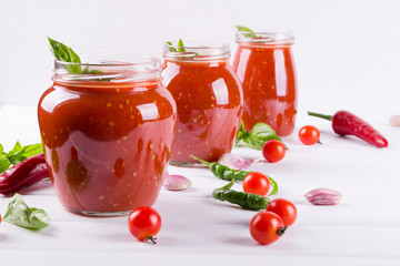 Fototapeta na wymiar Tomato ketchup sauce with cherry tomatoes and red hot chili peppers, garlic and herbs in a glass jar on white background. Homemade tomato sauce and fresh tomatoes