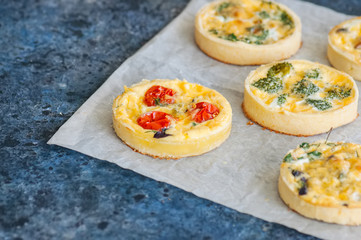 Set of savory mini tarts on a baking paper. Vegetable quiches with tomatoes, mushrooms, herbs, broccoly. Blue stone background. Close up and copy space.