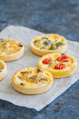 Set of savory mini tarts on a baking paper. Vegetable quiches with tomatoes, mushrooms, herbs, broccoly. Blue stone background. Close up and copy space.