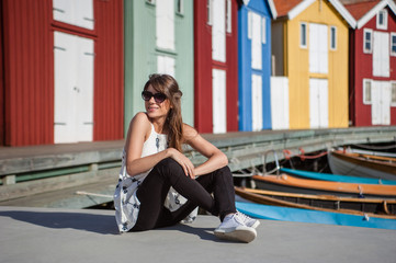 Obraz premium Young and happy tourist woman sitting in the harbor in Smogen town, Sweden. Beautiful traditional colorful wood houses on the background.