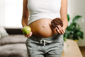 Pregnancy healthy diet and gluttony concept. Woman holding apple and chocolate cookie at home.