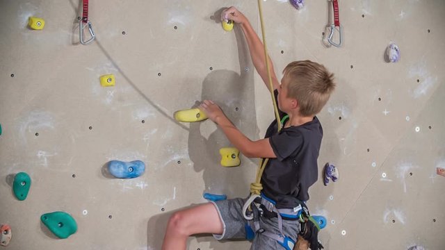 A young boy is very skilful at climbing. He is fastened with a safety rope so that he doesn't fall off. They're having PE in a school gym.