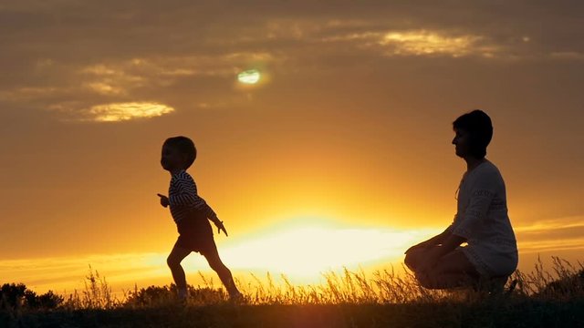 a silhouette of a happy young boy child running into the arms of his loving mother for a hug, in front of the sunset in the sky on a summer day.