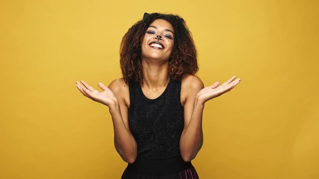 Tricky funny woman in haloween cat costume holding her heand together and smiling angrily to camera isolated over yellow 