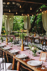 Wedding.Banquet.The festive table for guests,decorated with a composition of flowers and greenery,there are candles,served with cutlery and crockery,covered with the tablecloth in wooden summer house.