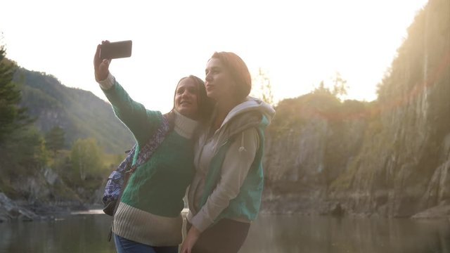 Young women is taking selfie on the phone against the background of big mountains and the green mountain river during sunset. 3840x2160