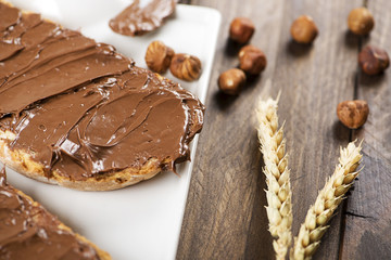 Chocolate spread on bread on a plate next to hazelnuts and wheat flower on brown wooden table. Food.