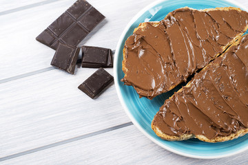 Chocolate spread on bread on a blue plate next to pieces of chocolate on wooden table. Copy space.