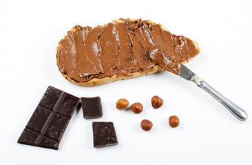 Chocolate spread on bread next to chocolate tablet, hazelnuts and knife on white background. Isolated.