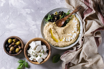 Homemade traditional spread hummus with chopping olives, oil and herbs on blue plate, served with olives, feta cheese, spoon, textile on gray texture background. Mediterranean snack. Flat lay - obrazy, fototapety, plakaty