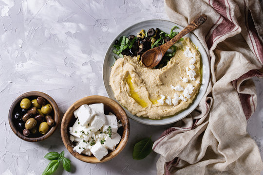 Homemade traditional spread hummus with chopping olives, oil and herbs on blue plate, served with olives, feta cheese, spoon, textile on gray texture background. Mediterranean snack. Flat lay