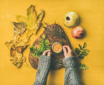 Autumn morning coffee concept. Flat-lay of woman' s hands in grey woolen sweater holding cup of espresso over mustard yellow background with dried fallen leaves and pomegranates around, top view