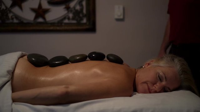Medium shot of a mature caucasian topless woman with stones in a row down her back.  Woman has smile on her face and is topless.