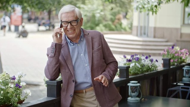 Joyous senior man in stylish suit and eyeglasses standing outdoors in summer cafe, smiling and talking on mobile phone