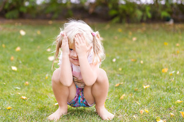 Portrait of cute adorable little blonde caucasian girl squatting on green grass lawn outdoors,...