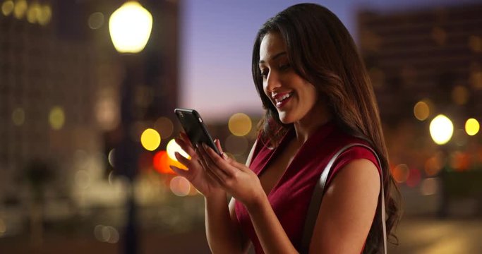 Attractive Latina female in sexy red dress taking a selfie with smart phone. Beautiful woman dressed for fun night in the city taking a picture of herself, smiling and laughing. 4k 