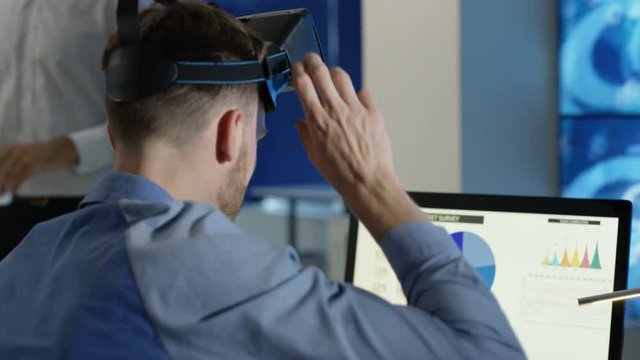  Businessman interacting with a virtual reality headset in futuristic office, computer screens showing financial information & digital code