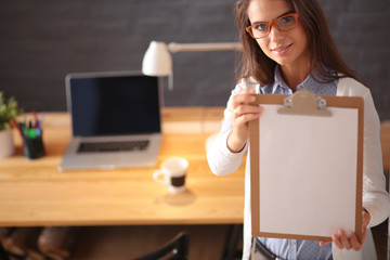 Young woman standing near desk with laptop holding folder and cup of coffee. Workplace. Business...