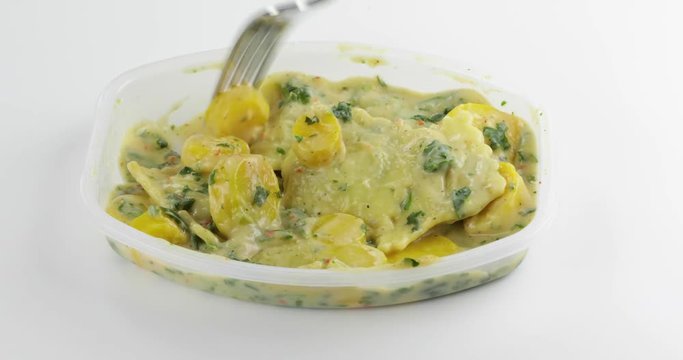 Video of a spinach ravioli TV dinner with vegetables in a microwavable tray being mixed with a fork then taking one bite of the pasta atop a white table top.