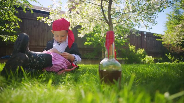 Little boy wearing pirate costume is looking at treasure map with spyglass. Pirate toy ship and bottle of rum on the lawn