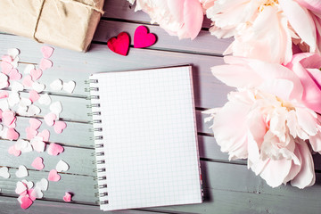 peonies flowers close-up with gift box, hearts and blank note book with copy space on wooden background. floral spring summer border template. mock-up sale, greeting and celebration card concept.